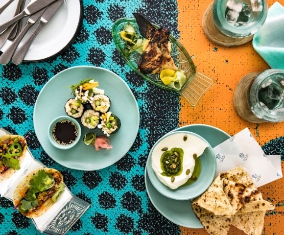 Overhead spread of sushi, avocado toast, dessert, drinks, and cutlery on a turquoise-and-orange patterned tablecloth; lively dining scene.