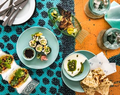 Overhead spread of sushi, avocado toast, dessert, drinks, and cutlery on a turquoise-and-orange patterned tablecloth; lively dining scene.