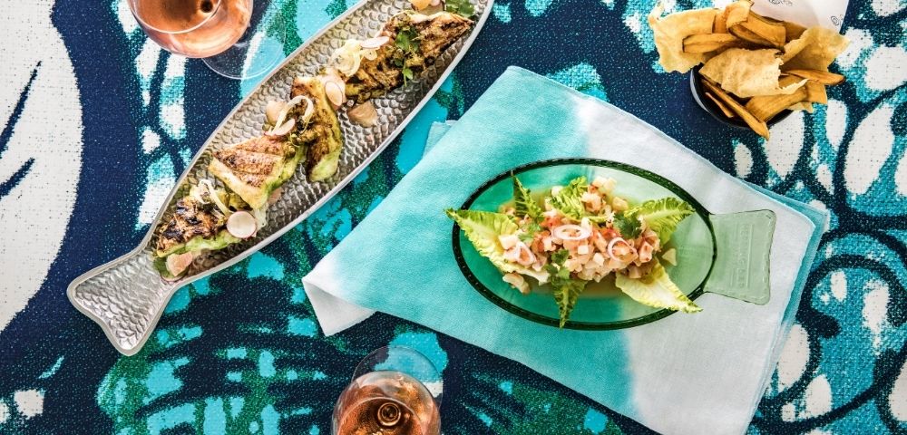 Overhead view of a colorful seafood spread: grilled fish platter, salad on a fish-shaped plate, fries, and two drinks on patterned fabric.