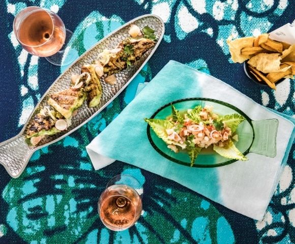 Overhead view of a colorful seafood spread: grilled fish platter, salad on a fish-shaped plate, fries, and two drinks on patterned fabric.