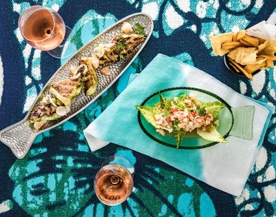 Overhead view of a colorful seafood spread: grilled fish platter, salad on a fish-shaped plate, fries, and two drinks on patterned fabric.
