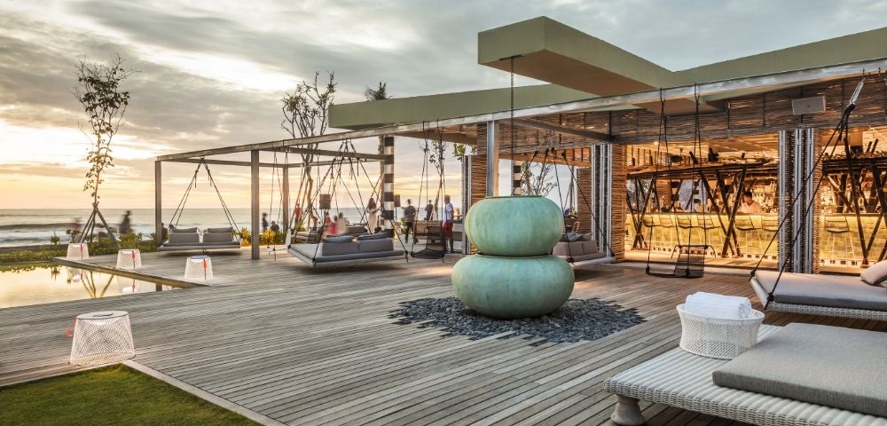 Contemporary seaside rooftop lounge at sunset, with swings, woven chairs, teal cushions, and a glass-walled bar overlooking calm water.