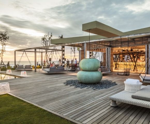 Contemporary seaside rooftop lounge at sunset, with swings, woven chairs, teal cushions, and a glass-walled bar overlooking calm water.