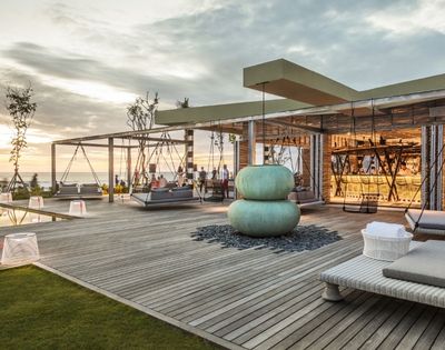 Contemporary seaside rooftop lounge at sunset, with swings, woven chairs, teal cushions, and a glass-walled bar overlooking calm water.