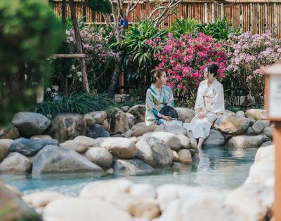 Two women in white robes sit beside a tranquil garden stream, surrounded by smooth stones, pink flowers, greenery, and a bamboo lantern.