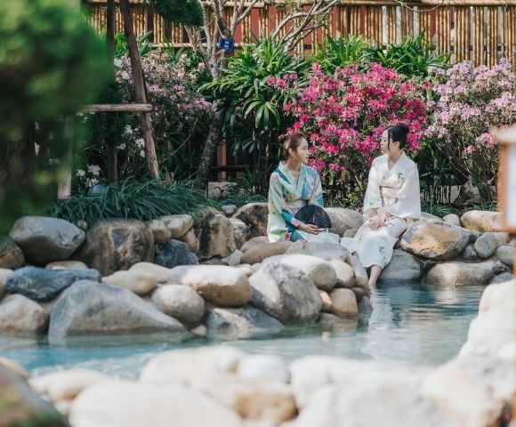 Two women in white robes sit beside a tranquil garden stream, surrounded by smooth stones, pink flowers, greenery, and a bamboo lantern.