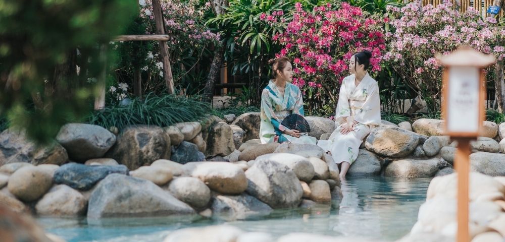 Two women in white robes sit beside a tranquil garden stream, surrounded by smooth stones, pink flowers, greenery, and a bamboo lantern.