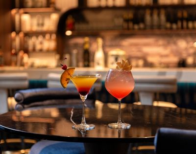 Two colorful cocktails garnished with citrus and fruit sit on a polished bar table, glowing warmly against a softly blurred, inviting lounge.
