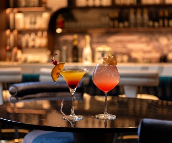 Two colorful cocktails garnished with citrus and fruit sit on a polished bar table, glowing warmly against a softly blurred, inviting lounge.