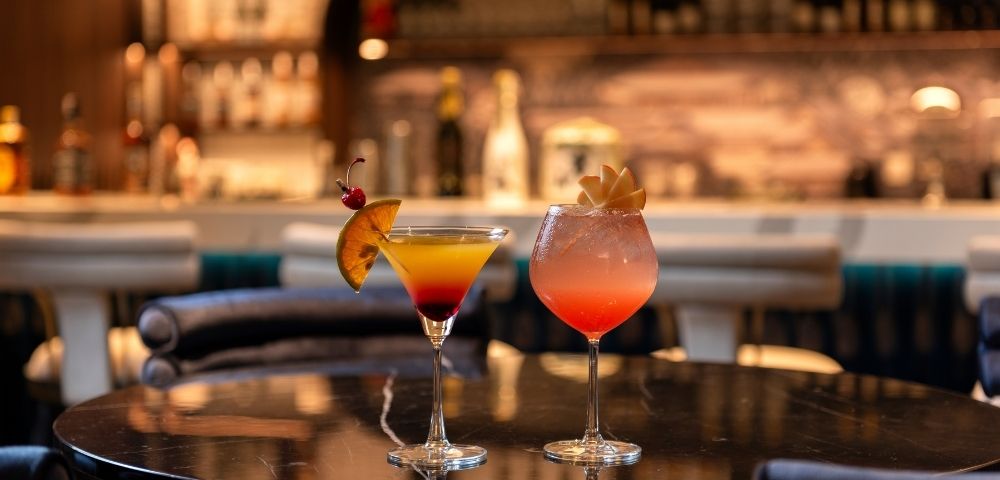 Two colorful cocktails garnished with citrus and fruit sit on a polished bar table, glowing warmly against a softly blurred, inviting lounge.