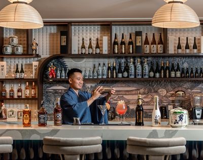 Bartender in a stylish Japanese bar prepares a cocktail behind a marble counter, surrounded by sake bottles, spirits, and warm pendant lighting.