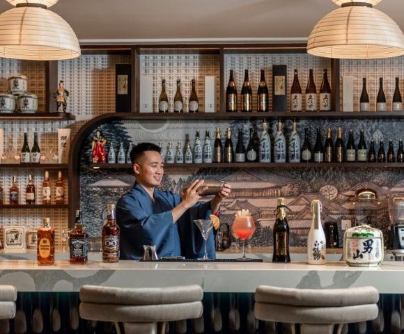 Bartender in a stylish Japanese bar prepares a cocktail behind a marble counter, surrounded by sake bottles, spirits, and warm pendant lighting.