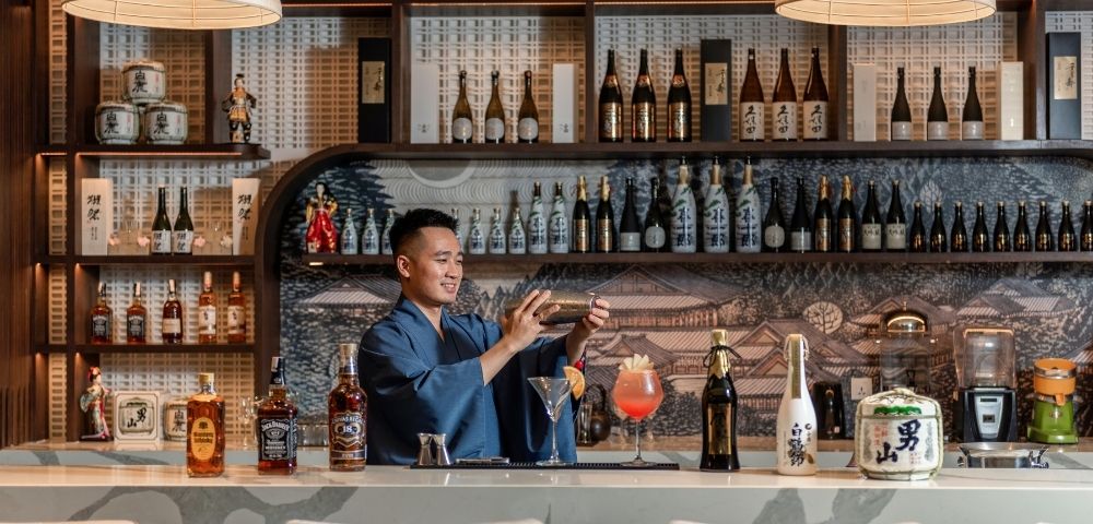 Bartender in a stylish Japanese bar prepares a cocktail behind a marble counter, surrounded by sake bottles, spirits, and warm pendant lighting.