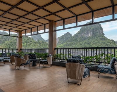 Covered open-air terrace with wicker chairs, sofas, plants, and wood flooring; dramatic green karst peaks create a serene atmosphere.
