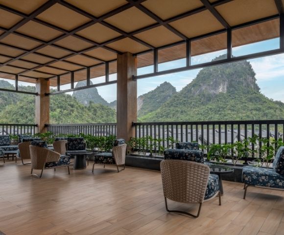 Covered open-air terrace with wicker chairs, sofas, plants, and wood flooring; dramatic green karst peaks create a serene atmosphere.
