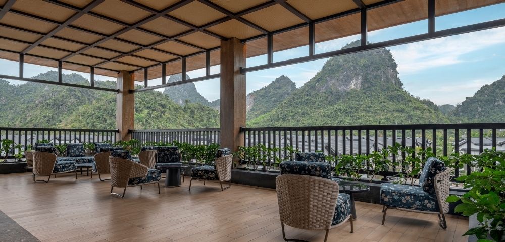 Covered open-air terrace with wicker chairs, sofas, plants, and wood flooring; dramatic green karst peaks create a serene atmosphere.