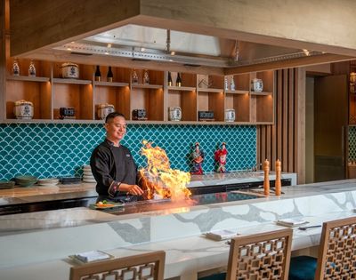 Chef theatrically cooks over a flaming teppanyaki grill in an elegant restaurant with turquoise tile, wood shelves, and warm lighting.