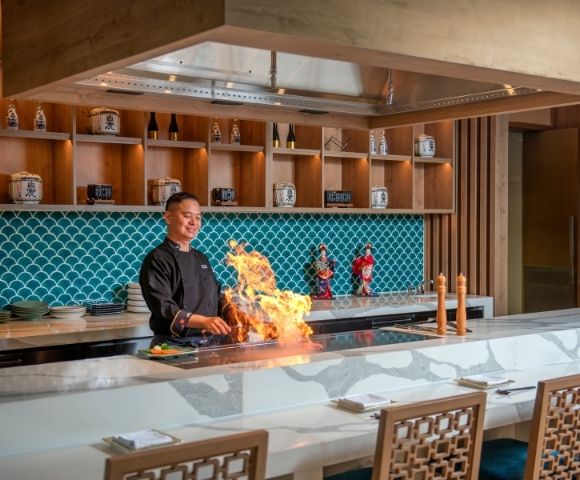 Chef theatrically cooks over a flaming teppanyaki grill in an elegant restaurant with turquoise tile, wood shelves, and warm lighting.