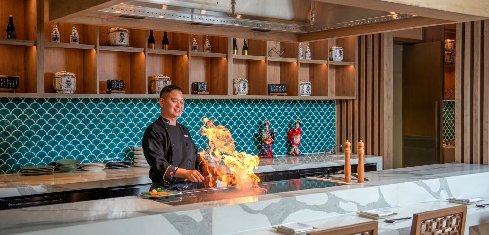 Chef theatrically cooks over a flaming teppanyaki grill in an elegant restaurant with turquoise tile, wood shelves, and warm lighting.