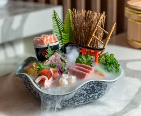 Japanese sushi boat platter with salmon, shrimp, tuna, roe, greens, and flowers, styled with bamboo, fern, and mist for a serene mood.