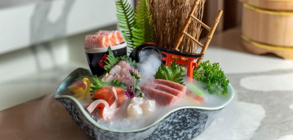 Japanese sushi boat platter with salmon, shrimp, tuna, roe, greens, and flowers, styled with bamboo, fern, and mist for a serene mood.
