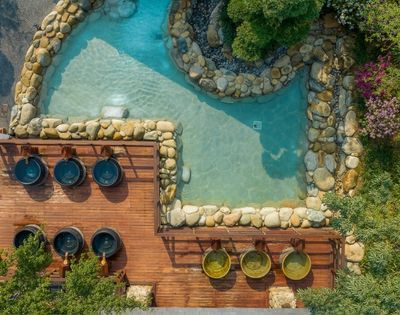 Aerial view of a serene spa area featuring a pond surrounded by rocks and plants, with wooden decks and small hot tubs.