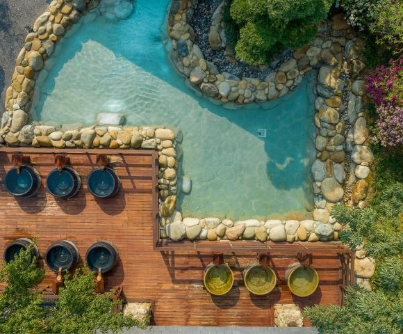 Aerial view of a serene spa area featuring a pond surrounded by rocks and plants, with wooden decks and small hot tubs.