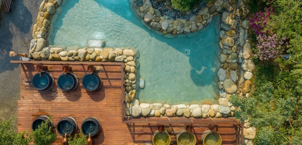 Aerial view of a serene spa area featuring a pond surrounded by rocks and plants, with wooden decks and small hot tubs.
