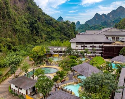 Aerial view of a resort surrounded by lush greenery and mountains, featuring multiple outdoor pools and pathways.