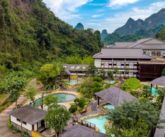 Aerial view of a resort surrounded by lush greenery and mountains, featuring multiple outdoor pools and pathways.