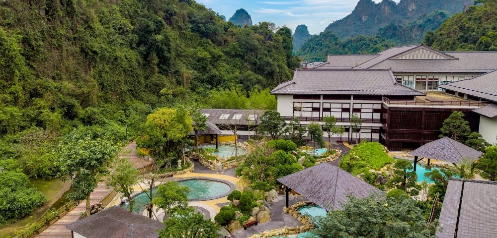 Aerial view of a resort surrounded by lush greenery and mountains, featuring multiple outdoor pools and pathways.