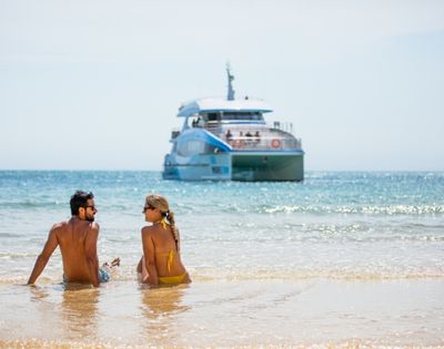 A couple sits on a sandy beach with waves lapping at their legs, looking out at a large, modern catamaran boat in the bright blue ocean under a clear sky.