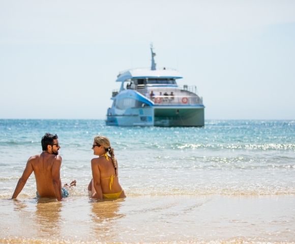 A couple sits on a sandy beach with waves lapping at their legs, looking out at a large, modern catamaran boat in the bright blue ocean under a clear sky.