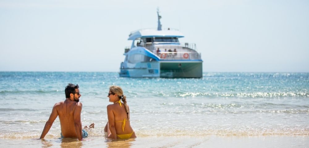 A couple sits on a sandy beach with waves lapping at their legs, looking out at a large, modern catamaran boat in the bright blue ocean under a clear sky.
