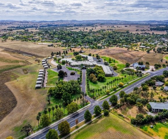 Aerial view of a sprawling resort complex with multiple buildings and palm tree-lined pathways, surrounded by residential areas, agricultural fields, and distant rolling hills under a cloudy sky.