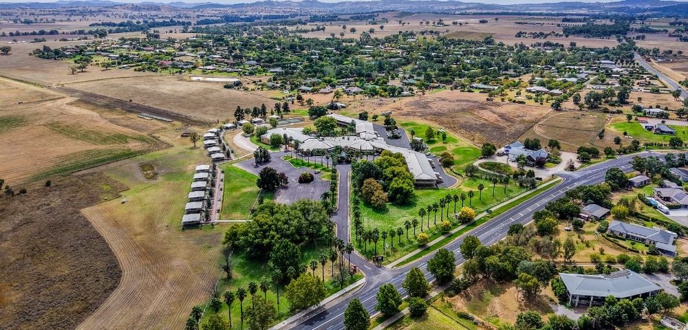 Aerial view of a sprawling resort complex with multiple buildings and palm tree-lined pathways, surrounded by residential areas, agricultural fields, and distant rolling hills under a cloudy sky.