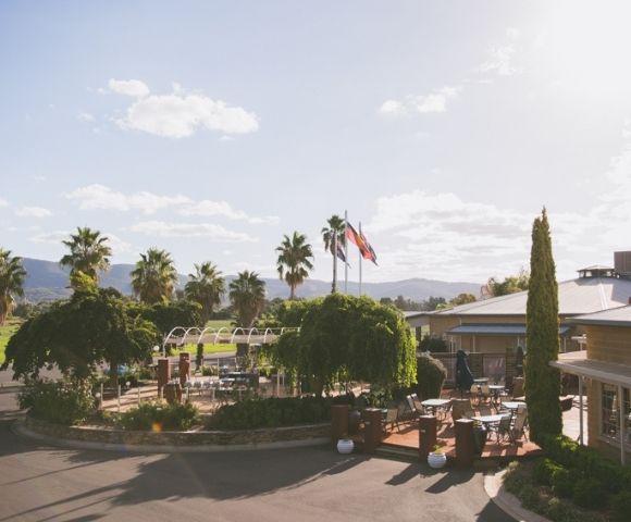 An elevated view of a resort with palm trees, an outdoor seating area with tables and chairs, flags flying, and buildings under a bright, sunny sky.