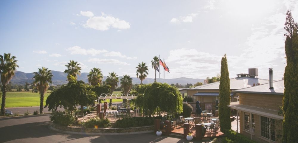 An elevated view of a resort with palm trees, an outdoor seating area with tables and chairs, flags flying, and buildings under a bright, sunny sky.