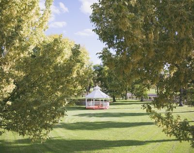 A white hexagonal gazebo with red trim sits on a green lawn surrounded by trees under a blue sky with scattered clouds.
