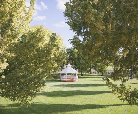 A white hexagonal gazebo with red trim sits on a green lawn surrounded by trees under a blue sky with scattered clouds.