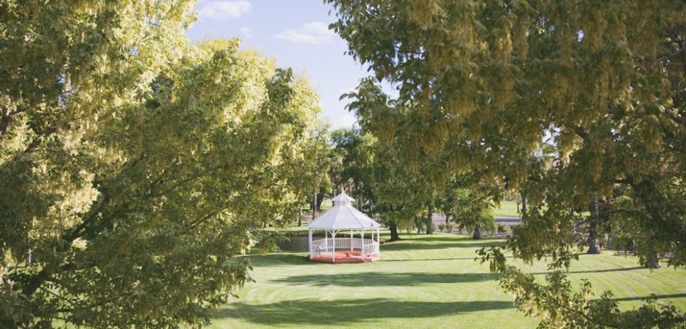 A white hexagonal gazebo with red trim sits on a green lawn surrounded by trees under a blue sky with scattered clouds.