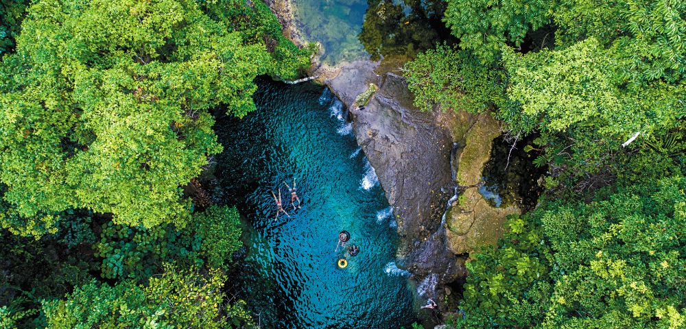 Aerial view of a natural blue pool surrounded by dense green forest with three people swimming and floating on inner tubes.