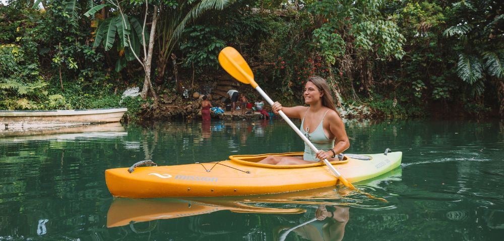 A woman paddles a bright orange kayak on calm green water surrounded by lush tropical trees and vegetation.