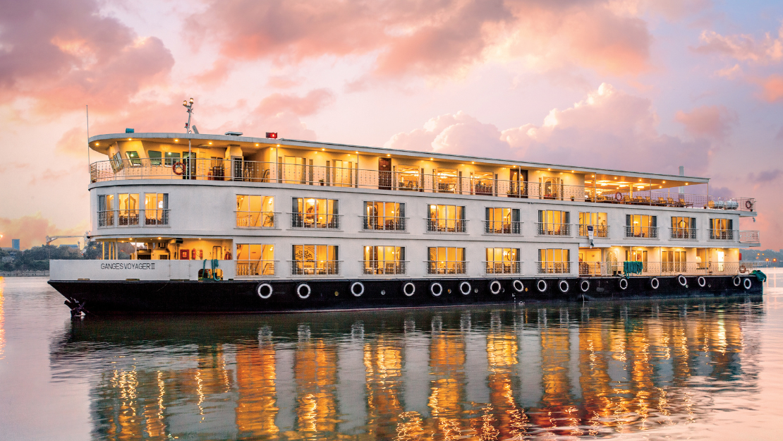 Luxury river cruise ship Ganges Voyager II illuminated at sunset, reflected on calm waters with a colorful sky backdrop.