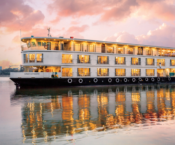 Luxury river cruise ship Ganges Voyager II illuminated at sunset, reflected on calm waters with a colorful sky backdrop.