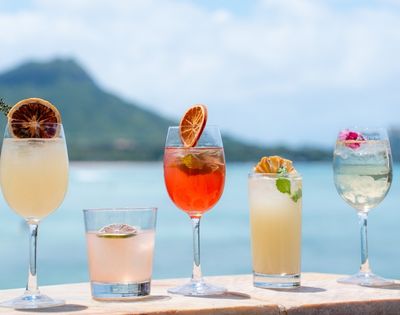 Five refreshing cocktails arranged on a ledge with a blurred tropical landscape and ocean in the background.