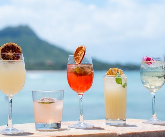 Five refreshing cocktails arranged on a ledge with a blurred tropical landscape and ocean in the background.