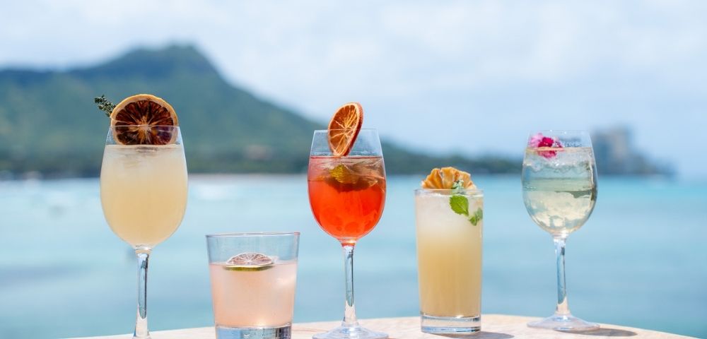 Five refreshing cocktails arranged on a ledge with a blurred tropical landscape and ocean in the background.