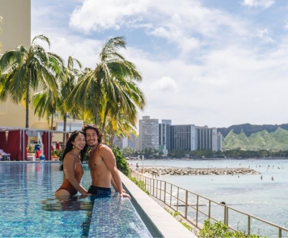 Couple in swimsuits embracing in an infinity pool with palm trees, hotels, and Diamond Head mountain in the background.