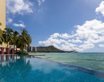 An infinity pool with blue water reflects palm trees and a resort building, overlooking a turquoise ocean with a distant mountain peak and scattered clouds.